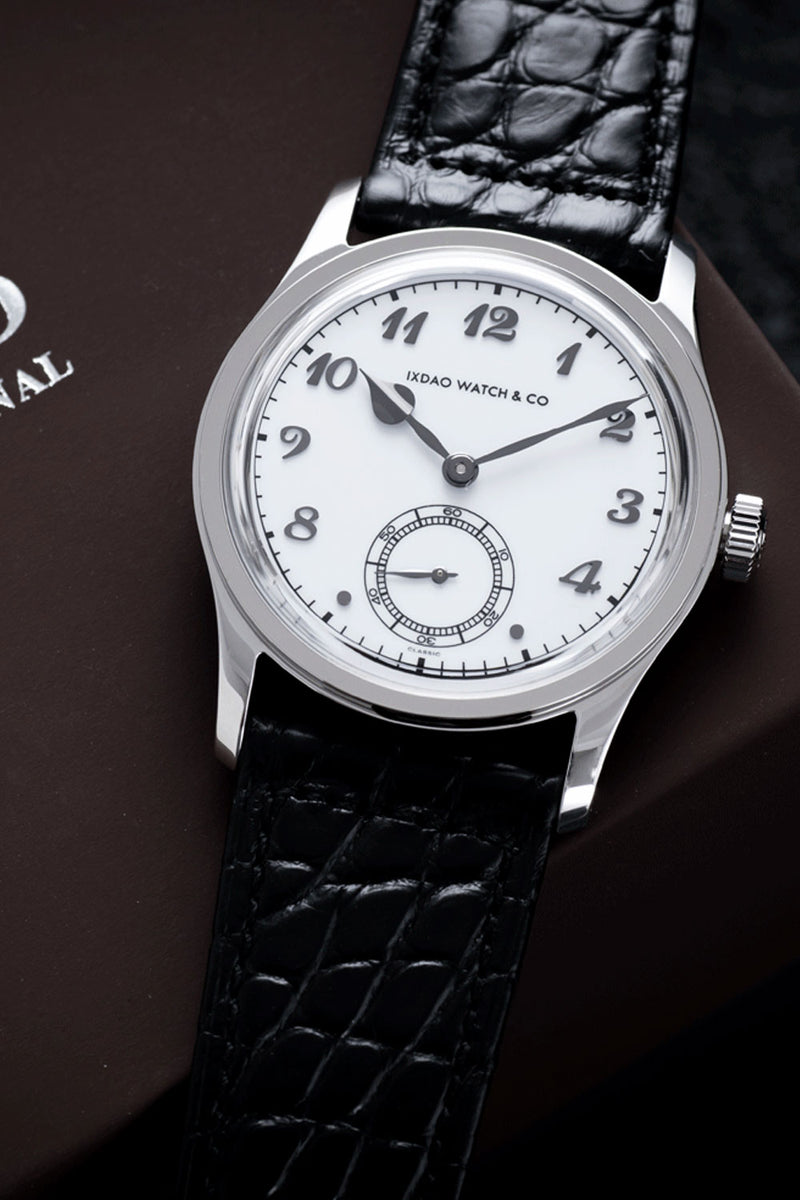 A close-up of a Iedo Watch & Co. wristwatch with a silver-toned round case, white dial with black Arabic numerals, and a black crocodile-embossed leather strap, resting on a dark brown surface.