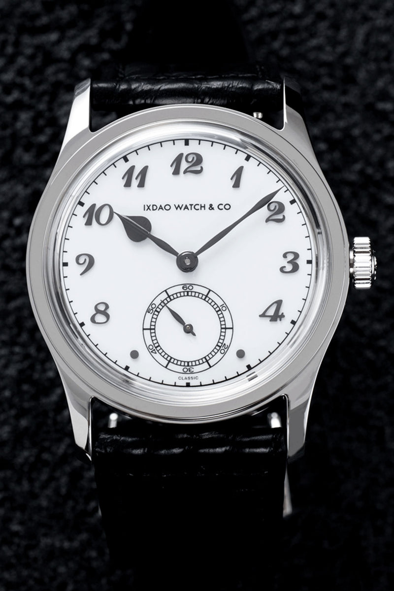 A close-up of a wristwatch with a silver-toned oval case, white dial, black Arabic numerals, black hands, and a black leather strap on a dark textured background.