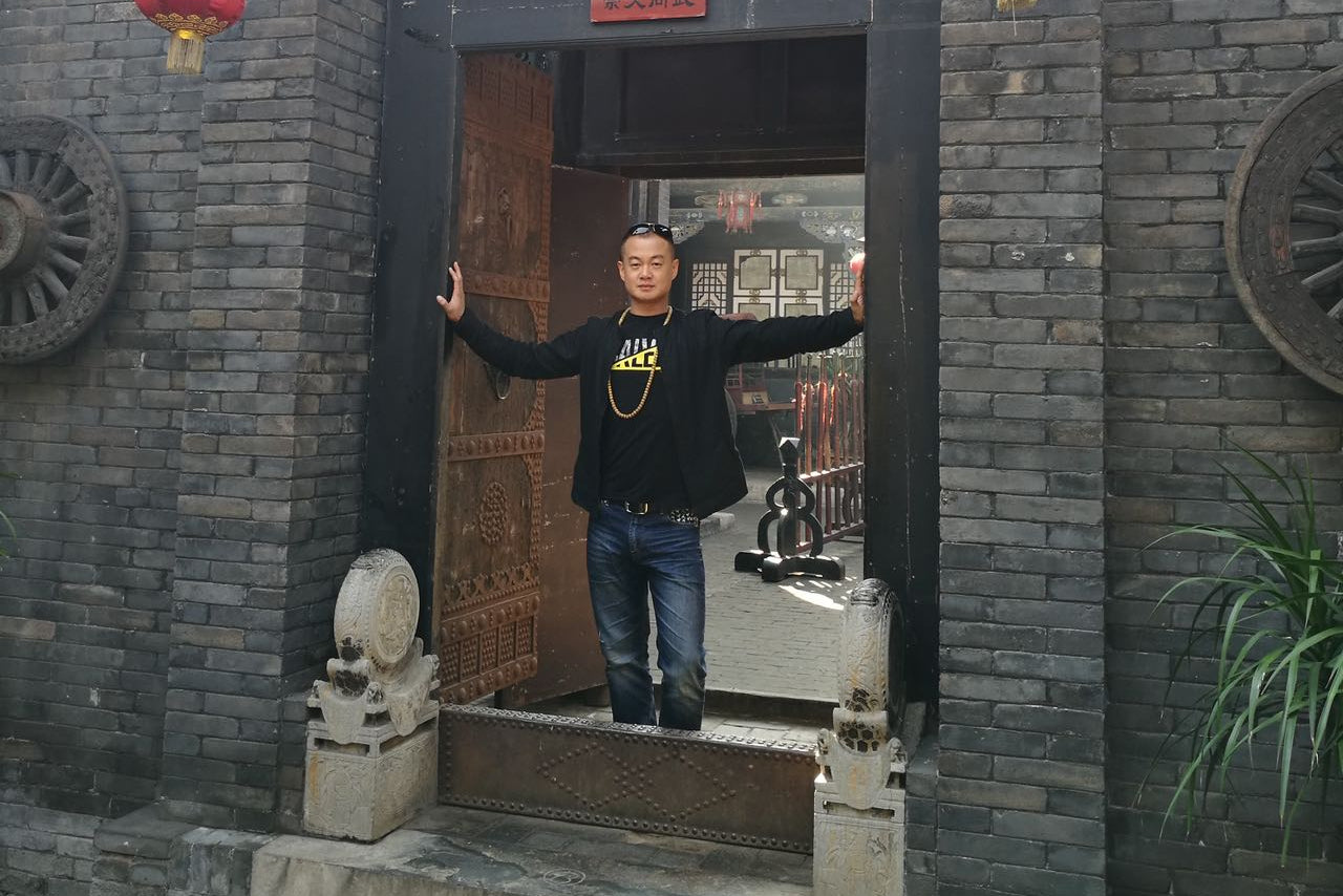 A man stands with arms outstretched in the doorway of a traditional Chinese brick building, wearing a black hoodie with a yellow graphic and blue jeans. Red lanterns hang on both sides of the dark wooden door, which has gold Chinese characters above him.