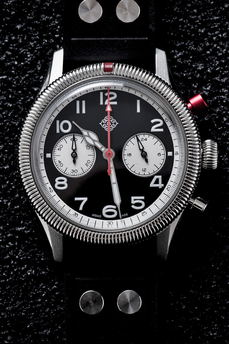 A wristwatch with a black dial, silver-tone bezel and case, white subdials, and a black strap, photographed against a dark textured background.