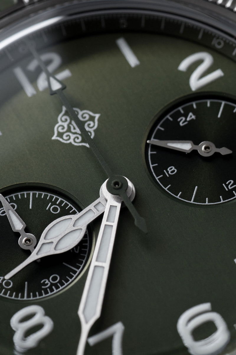 Close-up of a dark green watch dial with white Arabic numerals, silver hands, and two black subdials with white markings.