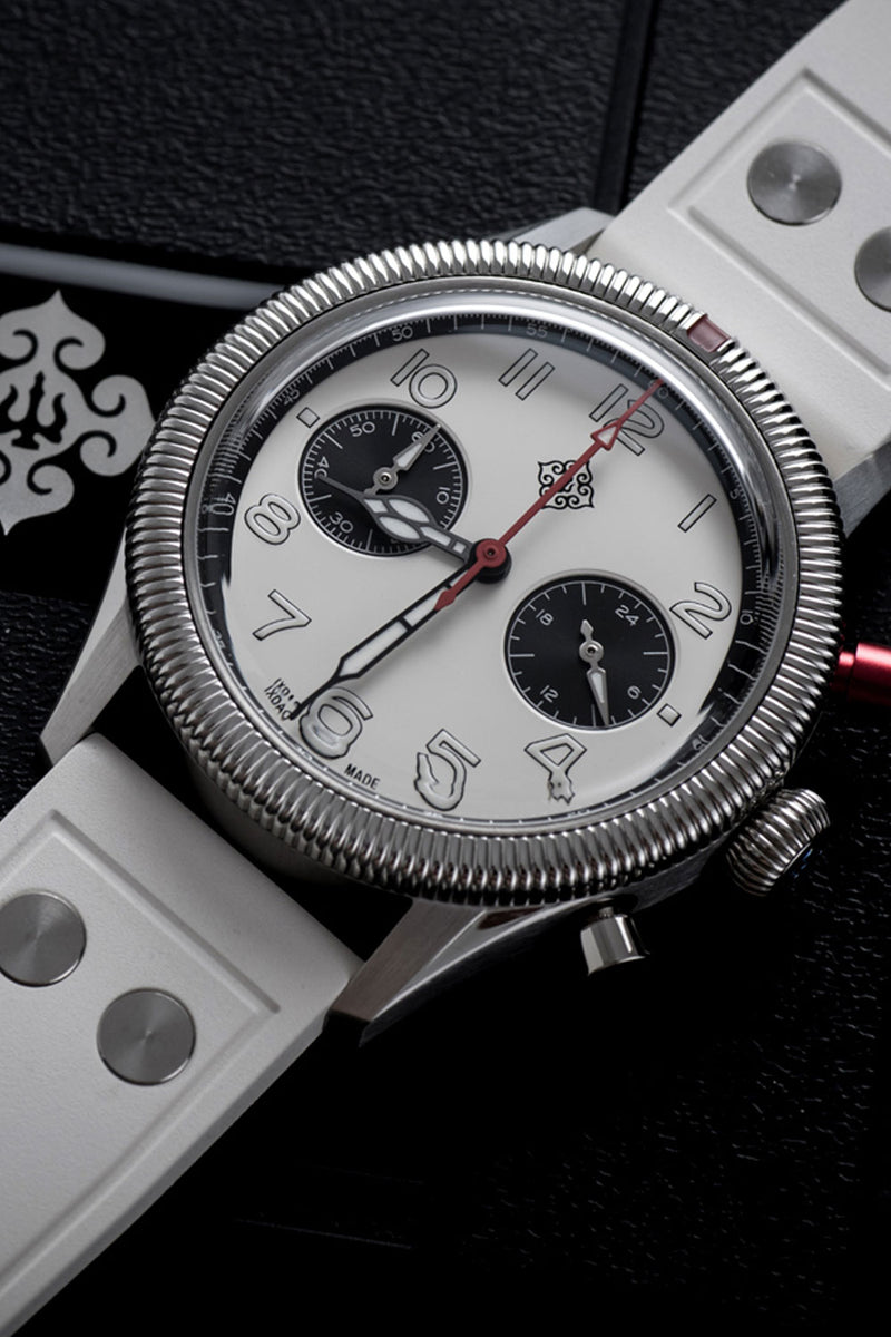 A close-up of a wristwatch with a silver-tone metal case, white dial, black subdials, and a white strap, photographed on a dark textured background.