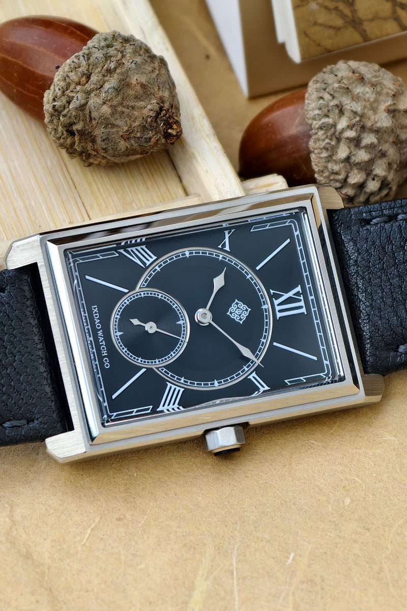 A rectangular silver-tone watch with a black dial, Roman numeral hour markers, a small subdial, and a black leather strap, styled on a tan surface with acorns and wood in the background.