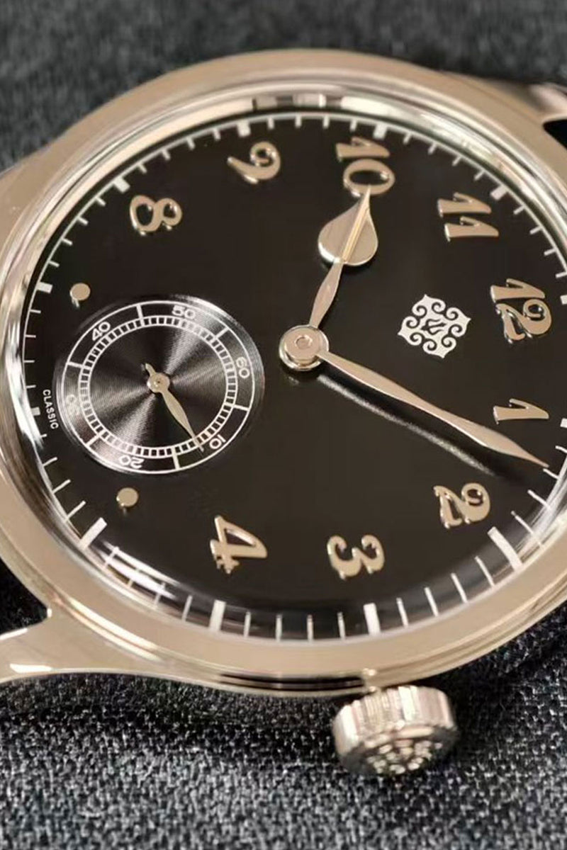Close-up of a wristwatch with a black dial, silver-tone case, and gold-toned numerals and hands, resting on a dark textured surface.
