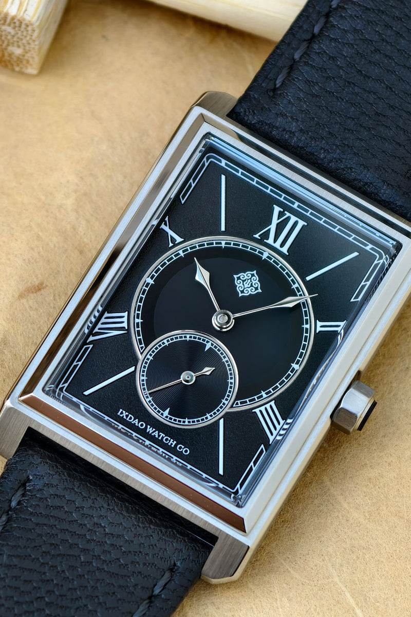 A close-up of a rectangular analog watch with a black dial, silver-toned case, and black textured leather strap, resting on a tan surface.
