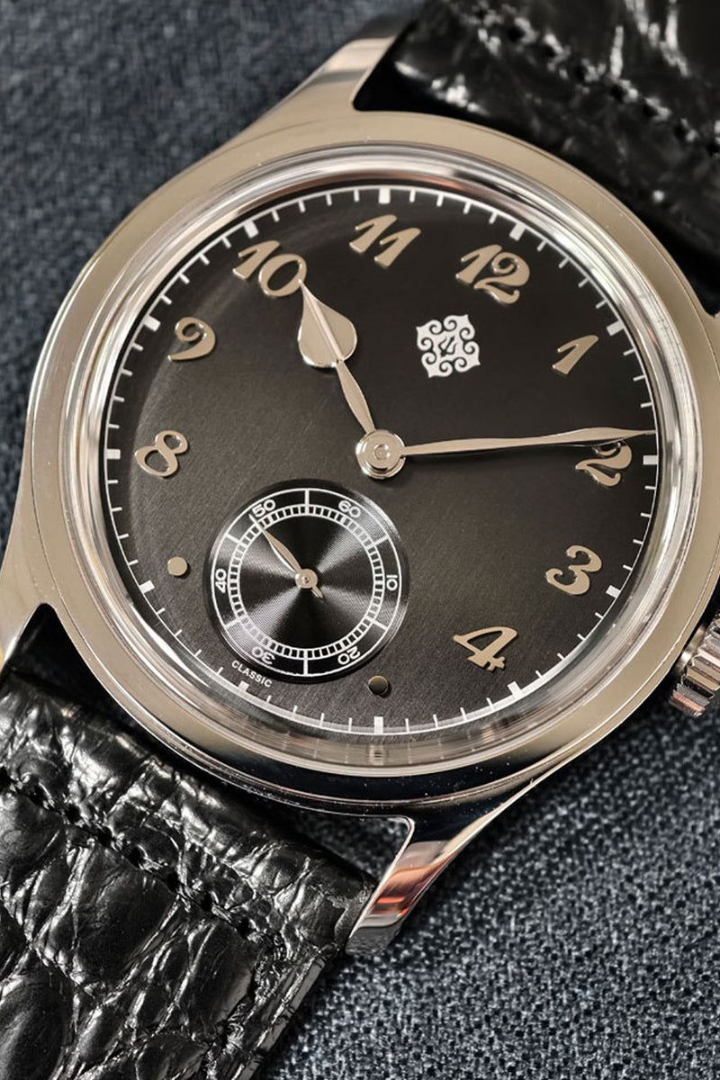 A close-up of a vintage-style wristwatch with a dark charcoal dial, gold-tone numerals, a small subdial at the 6 o'clock position, and a polished silver-tone case. The watch is fitted with a textured black leather strap and is photographed against a dark gray textured background.