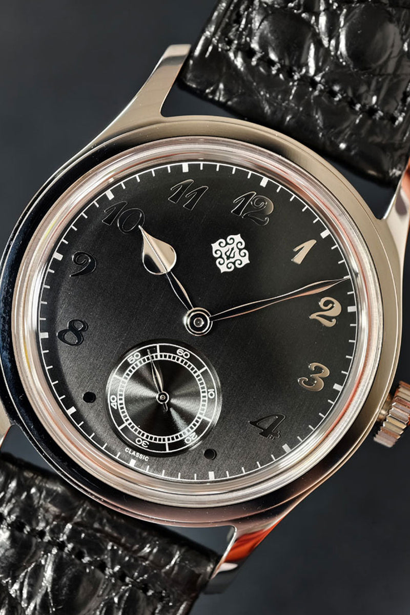 Close-up of a luxury wristwatch with a black dial, gold-tone numerals and hands, a small seconds subdial, and a black textured alligator leather strap.