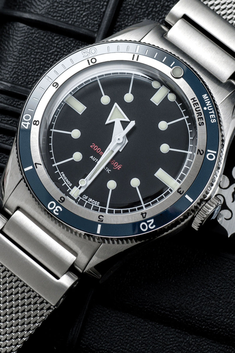 A close-up of a stainless steel wristwatch with a black dial, blue bezel, and silver mesh bracelet, resting on a dark textured surface.