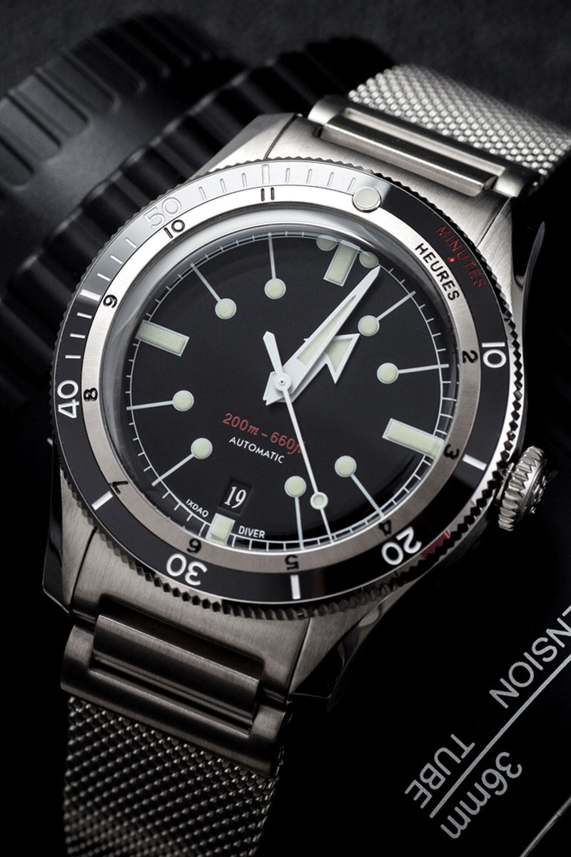 A close-up of a stainless steel wristwatch with a black dial, black rotating bezel, and matching black mesh strap, photographed on a dark textured background.