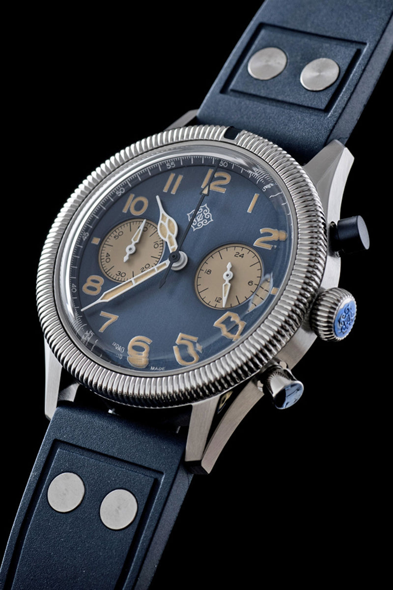 A close-up of a wristwatch with a dark blue leather strap, a silver-toned metal case, a blue dial, and beige subdials, shown against a solid black background.