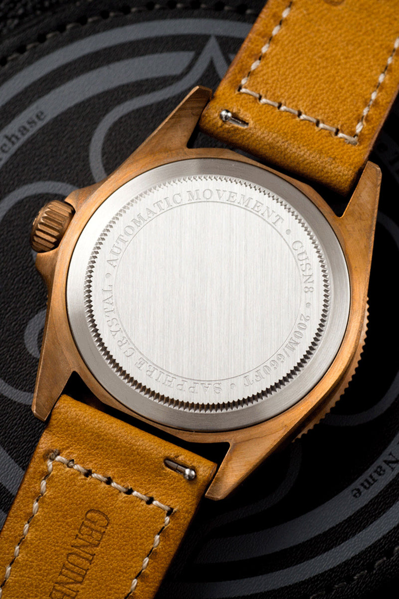 Close-up of the back of a wristwatch with a brushed silver case back, a copper-toned crown, and a tan leather strap on a dark patterned surface.