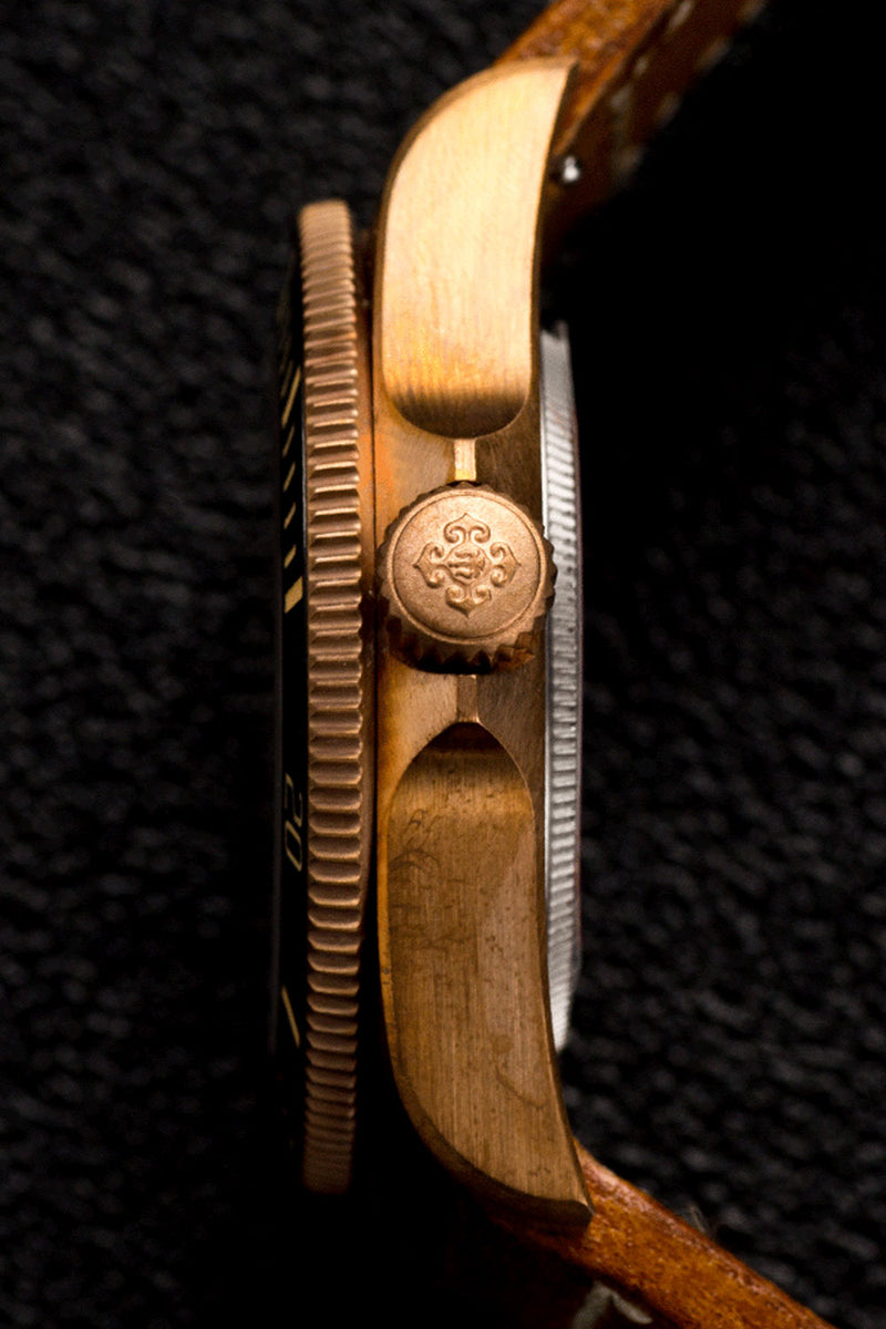 Close-up side view of a vintage wristwatch with a warm gold-toned metal case, a textured dark dial, a silver-toned inner ring, and a brown leather strap on a dark textured background.