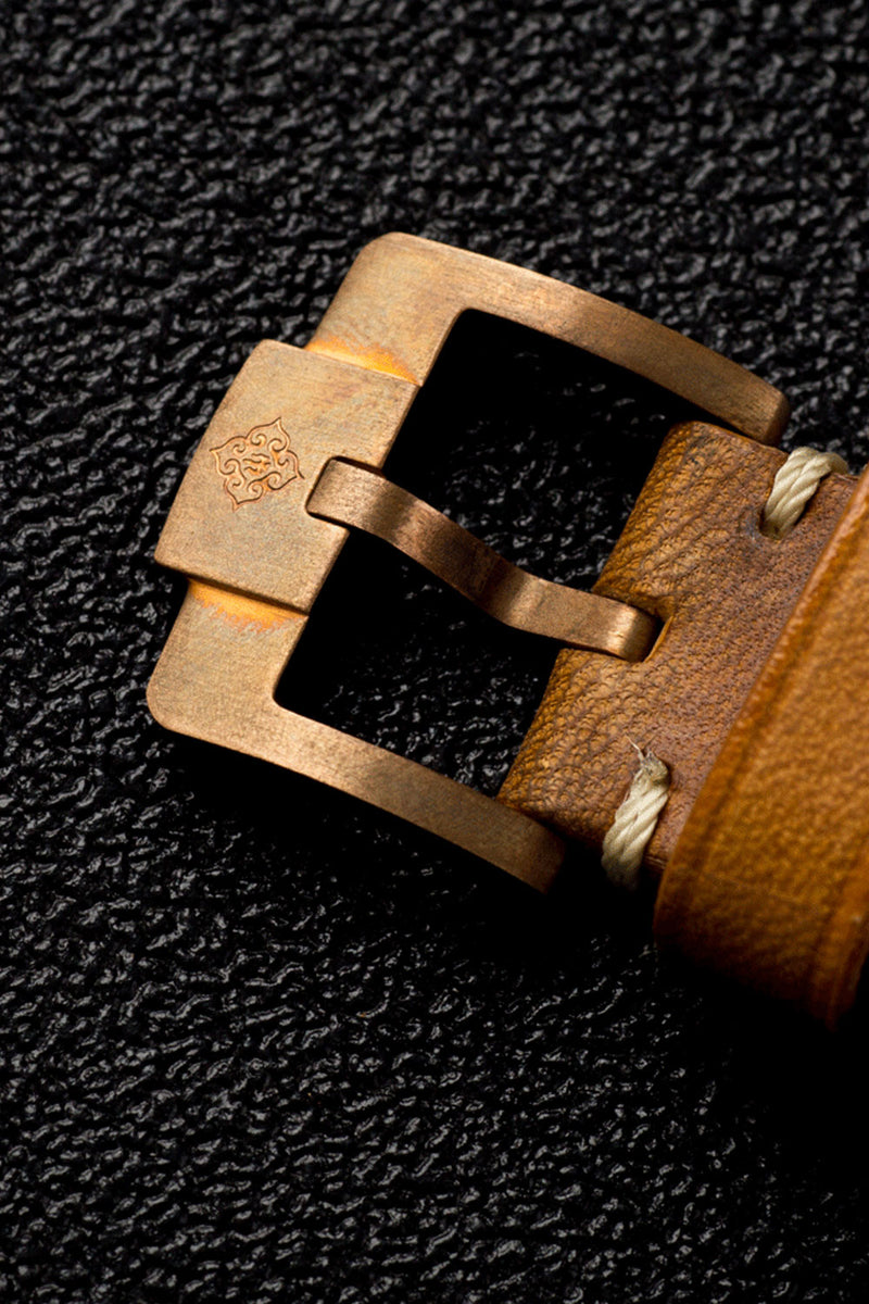 Close-up of a watch strap buckle with a warm brass-toned metal buckle and a brown leather strap on a dark textured background.