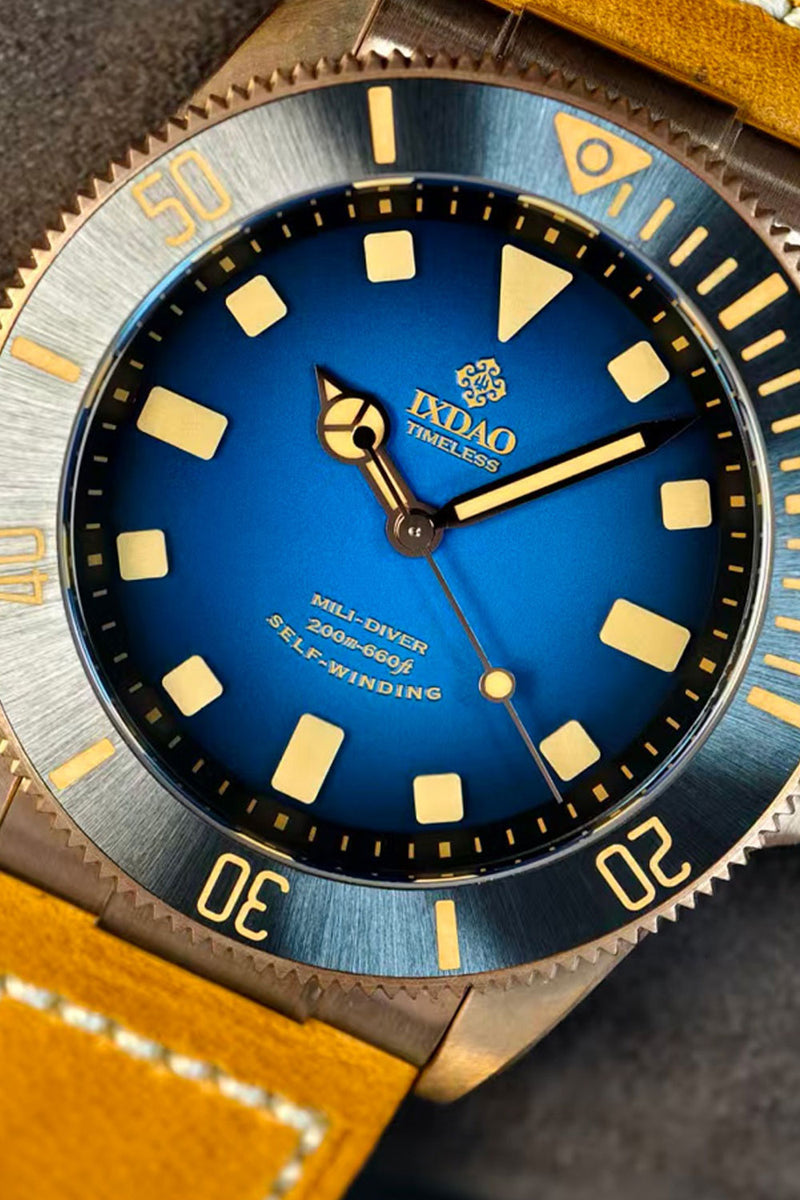 Close-up of an IDAO stainless steel dive watch with a blue dial, gold-tone markers and hands, and a textured blue and silver bezel. A yellow leather strap is partially visible.