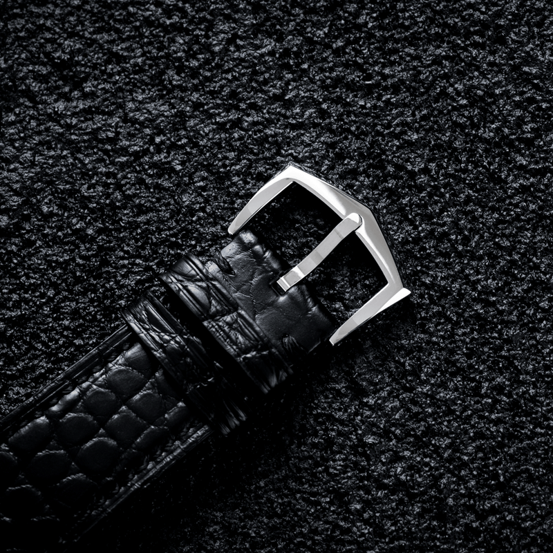 A close-up of a black textured leather watch strap with a silver-toned metal buckle, laid diagonally on a dark textured background.