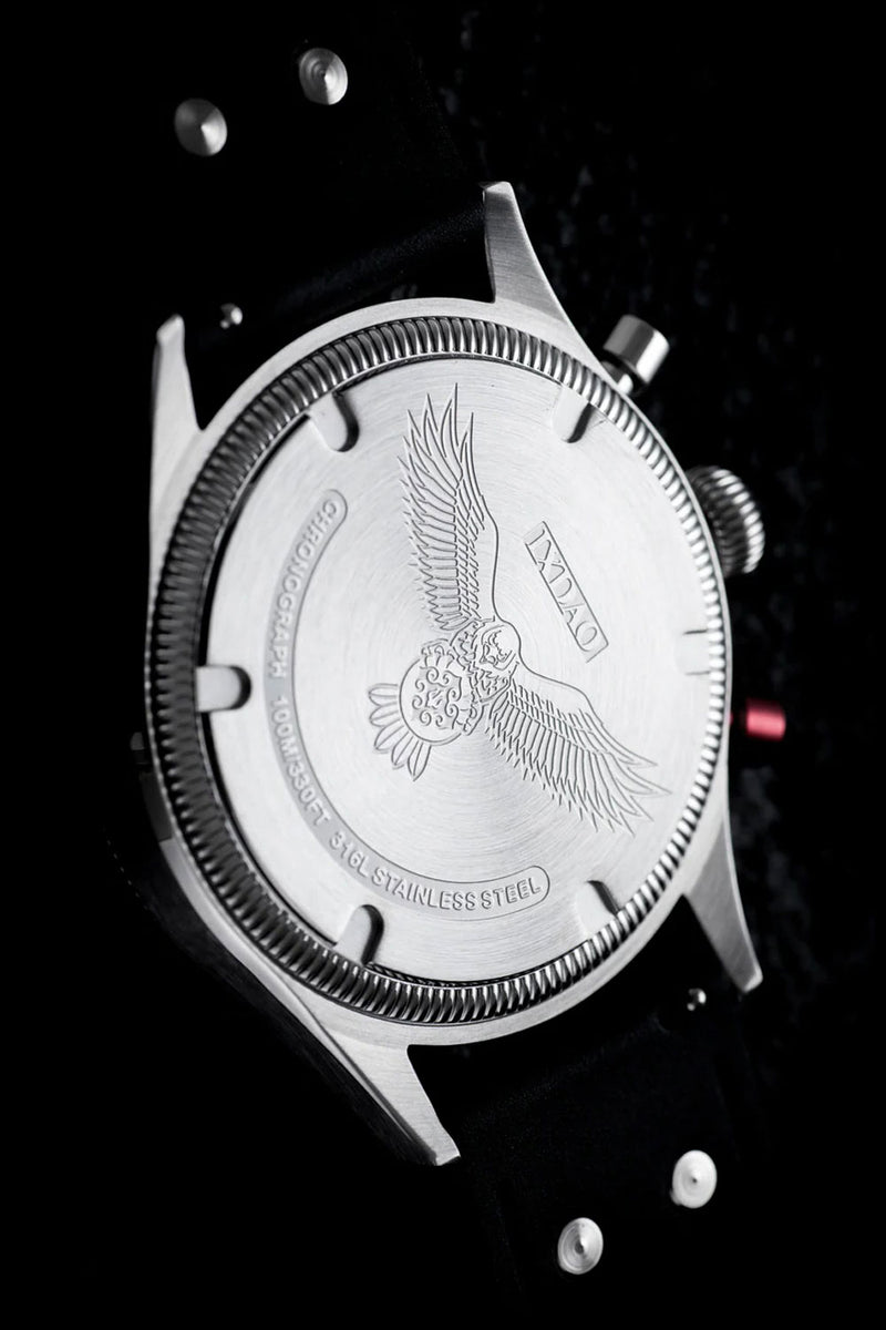 Close-up of the back of a stainless steel wristwatch with an engraved eagle motif and the brand name 'ANNU' on a black strap against a dark background.