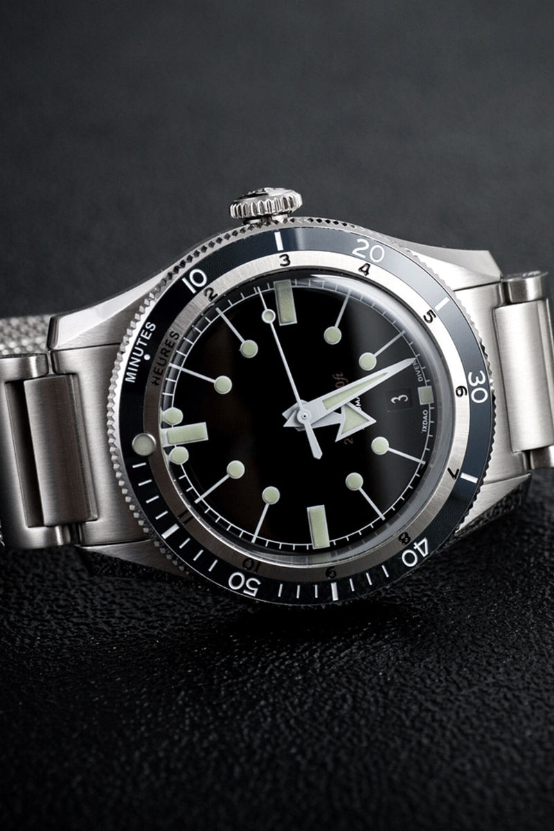 A stainless steel wristwatch with a black dial, black rotating bezel, and luminous silver-tone markers and hands, resting on a dark textured surface.