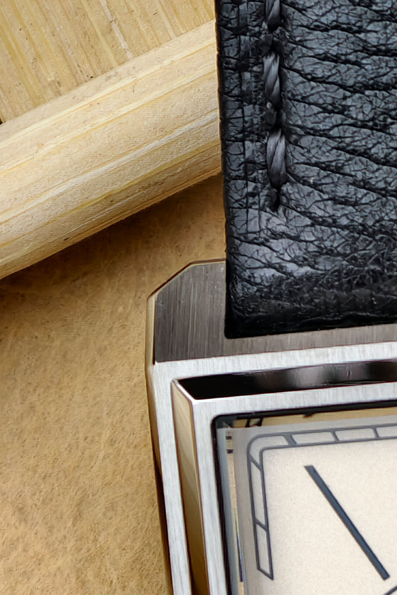 Close-up detail of a watch showing a black textured leather strap, brushed metal case, and cream-colored dial.