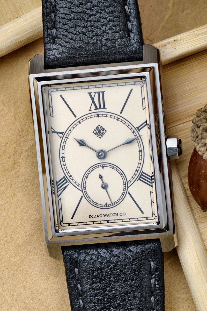 A rectangular stainless steel wristwatch with a cream-colored dial, Roman numeral hour markers, a small subdial, and a textured black leather strap, displayed on a wooden surface.