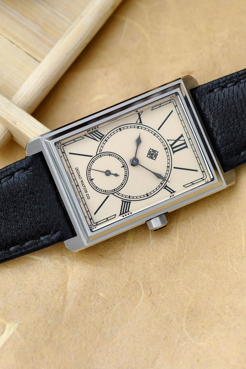 A rectangular silver-tone wristwatch with a cream dial, Roman numeral hour markers, a small subdial, and a black leather strap, photographed on a warm beige textured surface with blurred wooden props in the background.