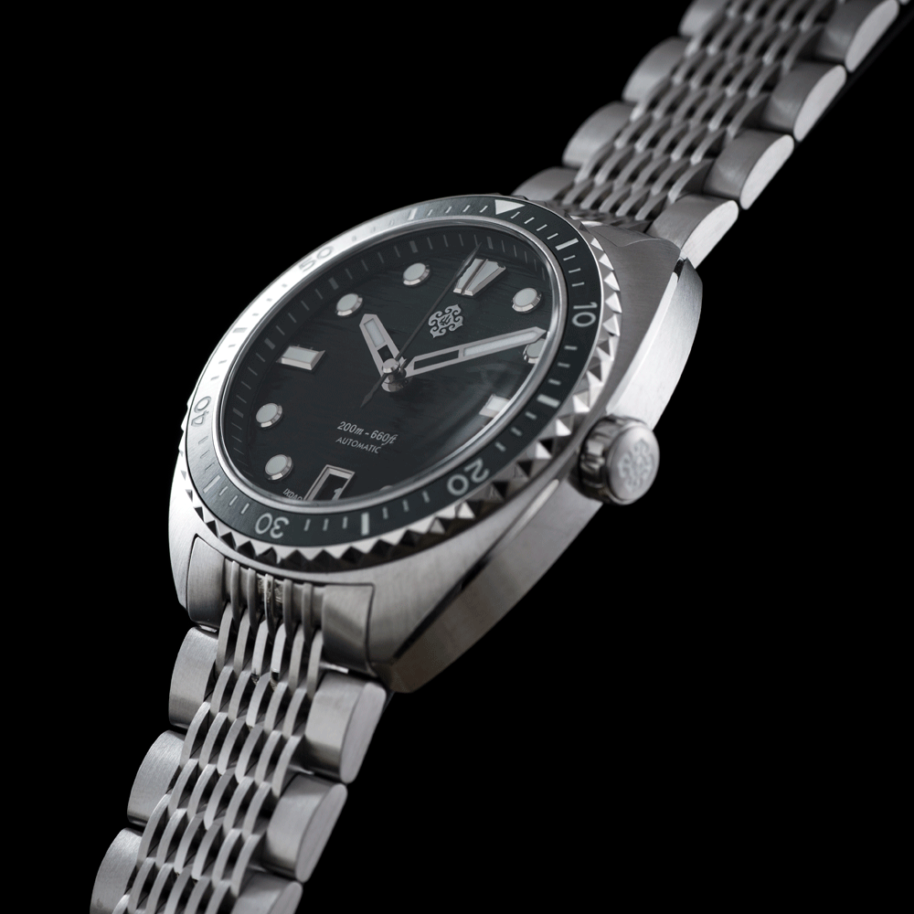 A stainless steel wristwatch with a black dial, black bezel, and matching steel link bracelet, photographed on a solid black background.