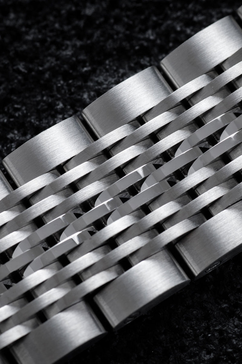 Close-up of a brushed stainless steel link bracelet with a woven or interlaced band design, shown against a dark textured background.