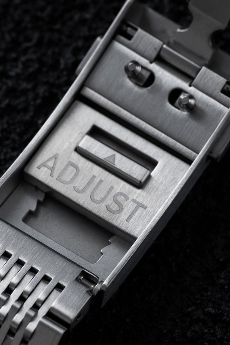Close-up of a brushed stainless steel watch band clasp with the word 'ADJUST' engraved on the front, set against a dark textured background.