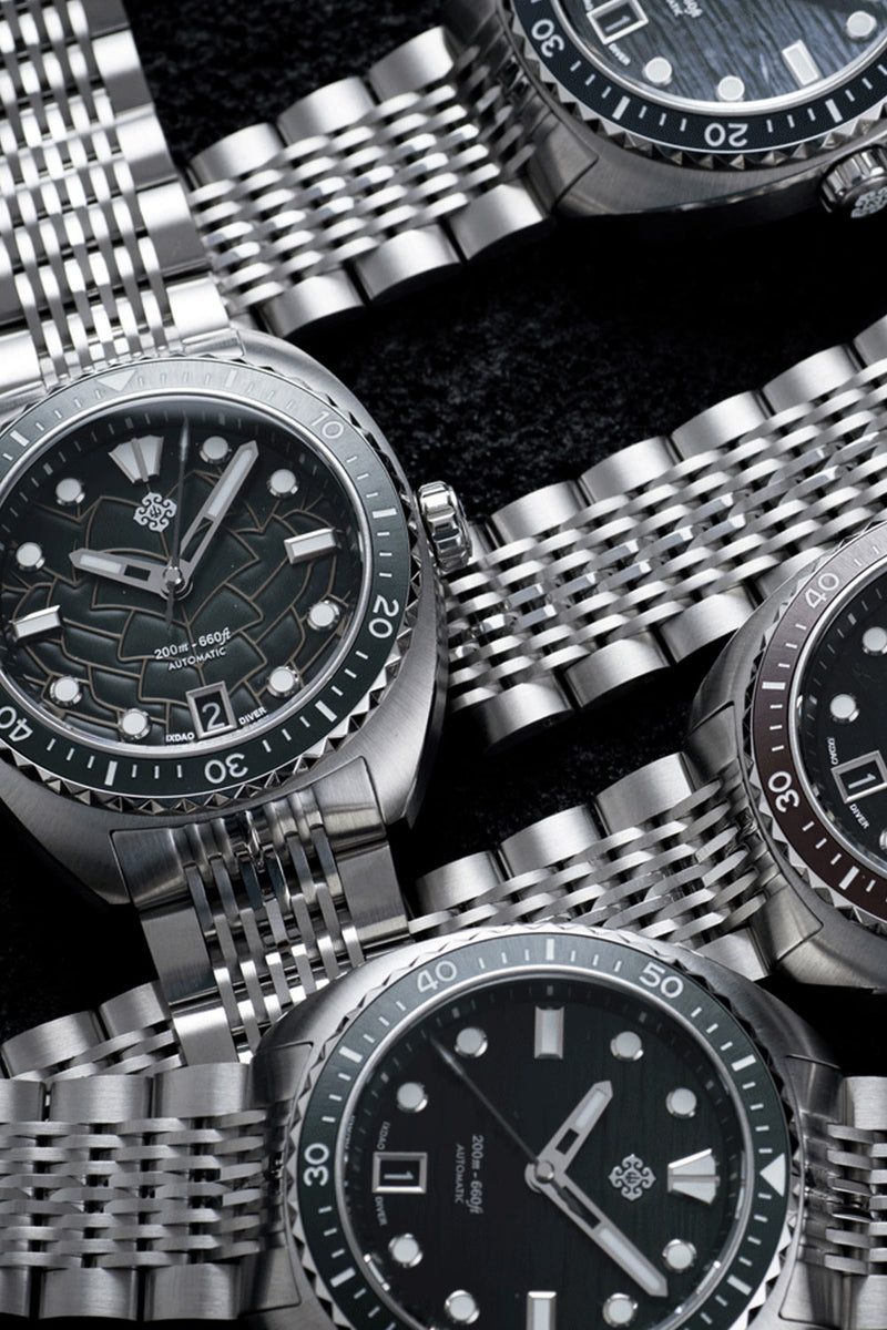 A close-up arrangement of stainless steel wristwatches with integrated metal bracelets and dark dials, photographed against a black textured background.