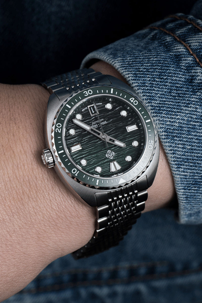 A close-up of a wrist wearing a silver-toned metal bracelet watch with a dark green textured dial and a green bezel, paired with a blue denim wristband.
