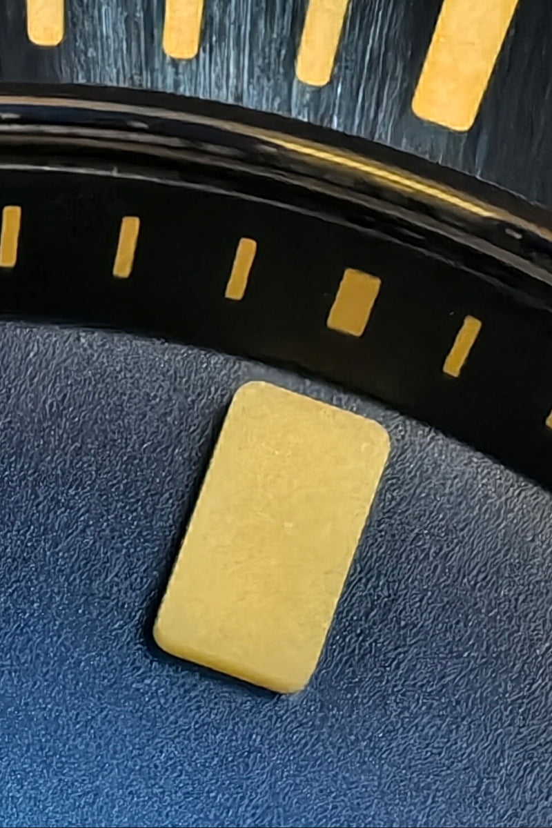 A small rectangular pale yellow stone or resin tag with rounded corners, placed on a textured dark blue surface with a black curved rim and gold markings in the background.