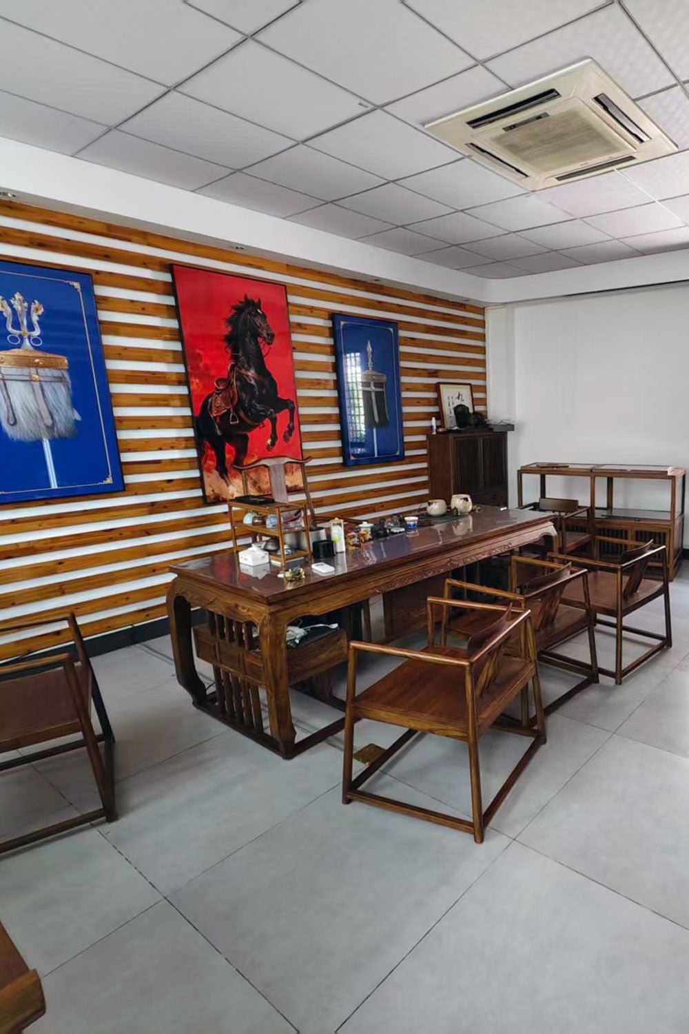 A traditional wooden tea table set with matching chairs in a modern office or showroom, featuring a wood-paneled wall with framed art and a red horse painting.