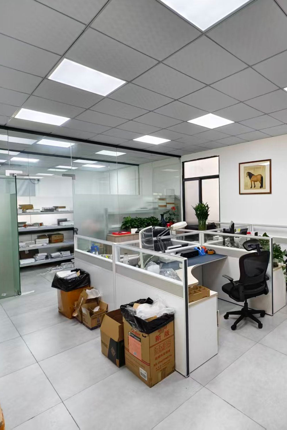 A modern office interior with white cubicles, a black office chair, a glass partition wall, tiled floors, and ceiling-mounted square lights. Several cardboard boxes and black trash bags are stacked on the floor near the desks.