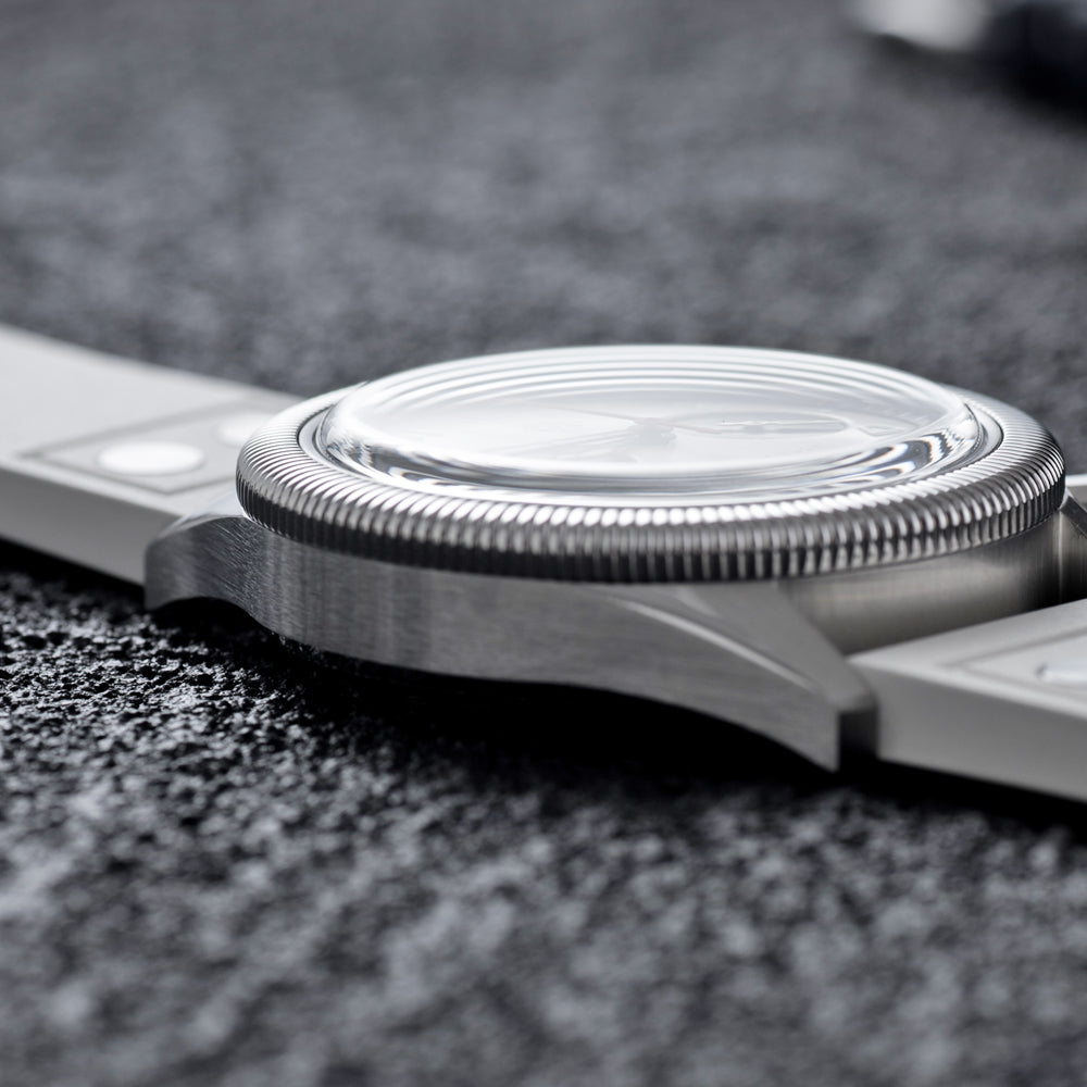 Close-up of a brushed silver metal watch case with a textured bezel and a clear crystal, resting on a dark textured surface.