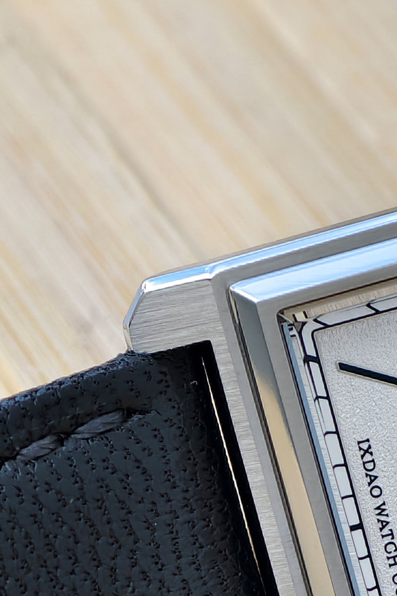 Close-up of a rectangular metal watch case with a black textured leather strap and a light dial, photographed on a light wood surface. The dial shows partial text reading 'IXDAO WATCH'.