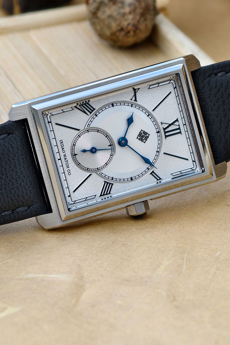 A rectangular silver-tone wristwatch with a white dial, Roman numeral hour markers, blue hands, and a black leather strap, resting on a light wood surface.