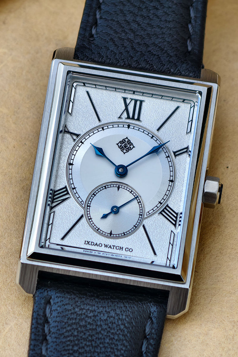 A close-up of a rectangular stainless steel watch with a silver dial, Roman numeral hour markers, blue hands, and a textured dark blue leather strap on a tan background.