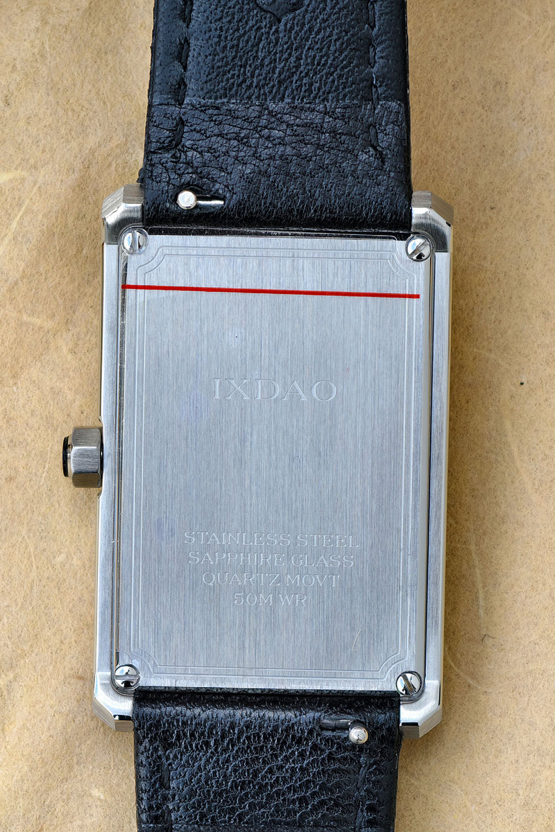 Close-up of the back of a rectangular stainless steel watch case with a black leather strap, photographed on a tan textured surface. The case shows engraved text including 'IXDAO' and 'STAINLESS STEEL SAPPHIRE GLASS'.