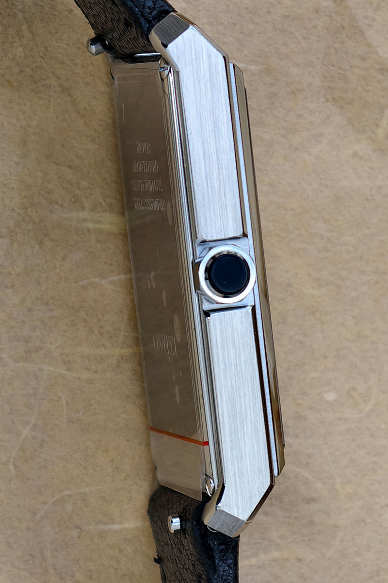 A close-up side view of a slim rectangular silver-toned metal watch with a dark circular bezel detail and a dark textured strap, photographed on a beige stone-like surface.