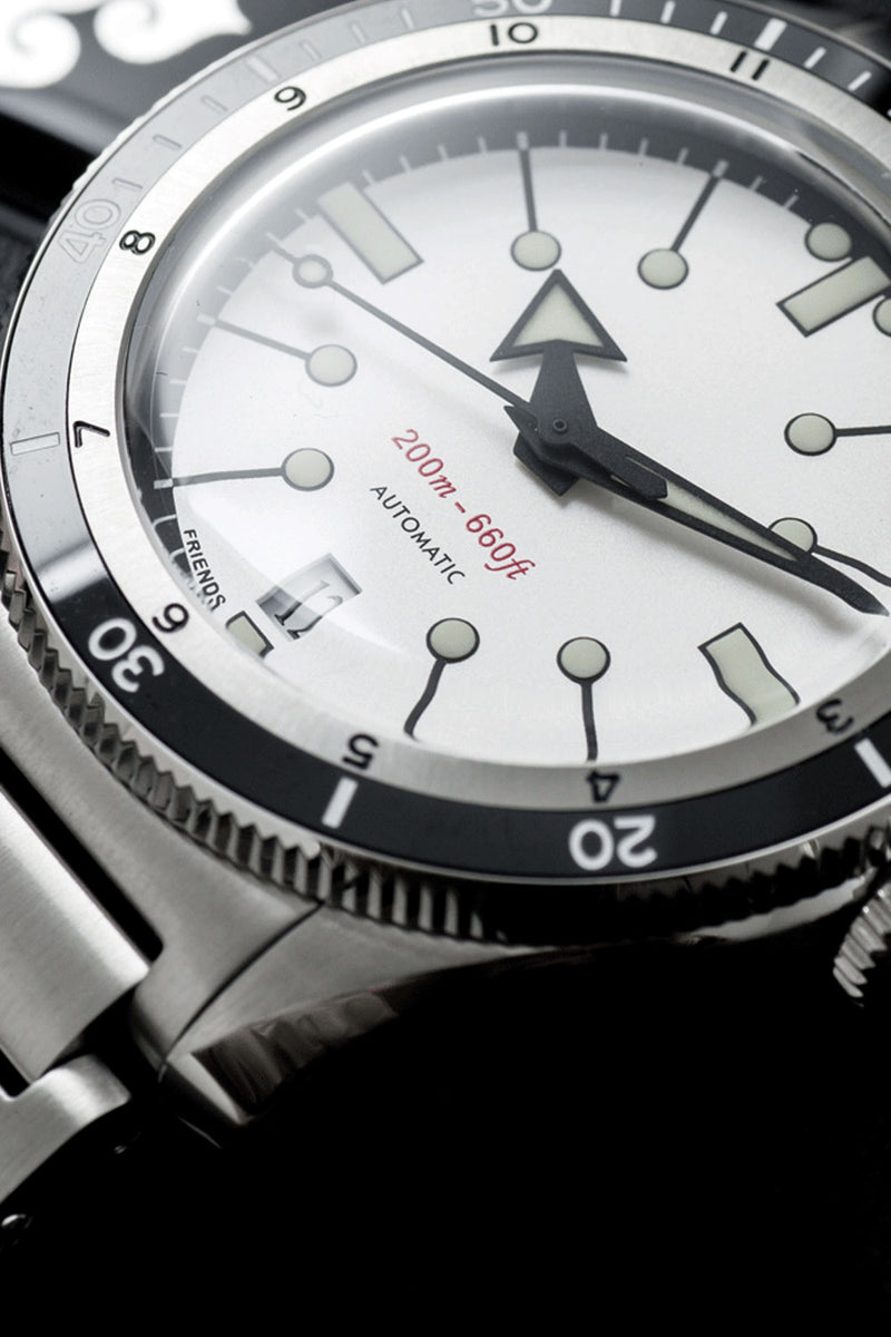 Close-up of a stainless steel wristwatch with a white dial, black bezel, and silver-tone metal bracelet against a dark background.