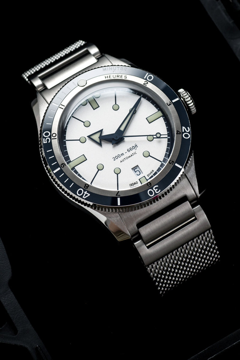 A Heuer wristwatch with a stainless steel mesh bracelet, featuring a white dial, a black rotating bezel, and silver-tone lugs, shown against a dark background.