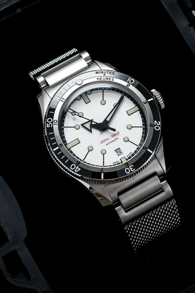 A stainless steel wristwatch with a silver mesh bracelet, featuring a white dial, black bezel, and green-luminescent hour markers, photographed against a dark background.
