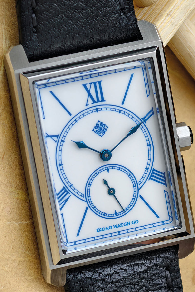 A close-up of a rectangular stainless steel wristwatch with a white dial, blue Roman numerals, blue hands, and a black textured leather strap, resting on a beige surface.