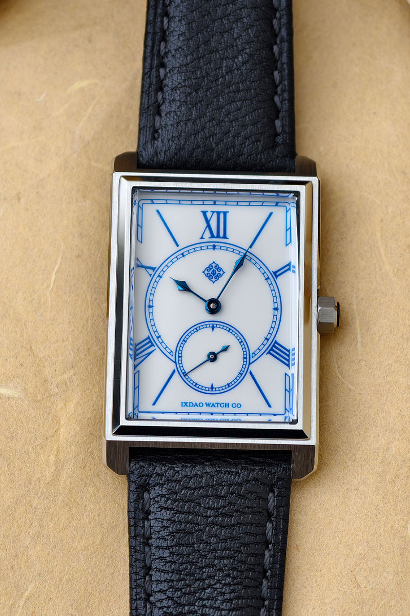 A rectangular analog watch with a white dial, blue Roman numerals, blue hands, and a blue inner subdial, housed in a silver-toned metal case with a black textured leather strap, shown on a beige textured background.