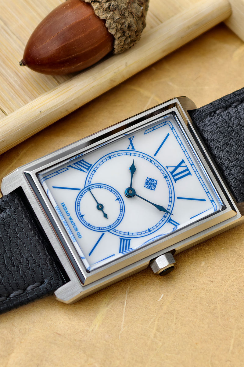 A rectangular analog watch with a white dial, blue Roman numerals, blue hands, and a silver-toned metal case, shown on a beige surface with a black textured strap. A wooden rod and acorn are partially visible in the background.