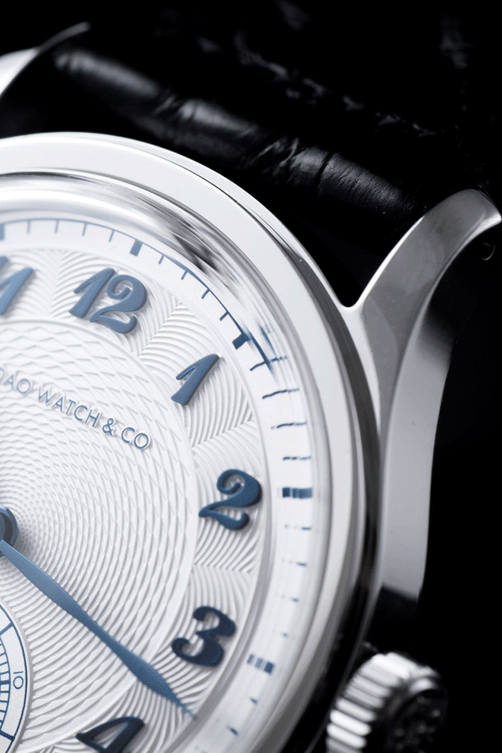 Close-up of a Bao Watch & Co. wristwatch featuring a white textured dial with blue Arabic numerals, a polished silver-tone case, and a black leather strap against a dark background.