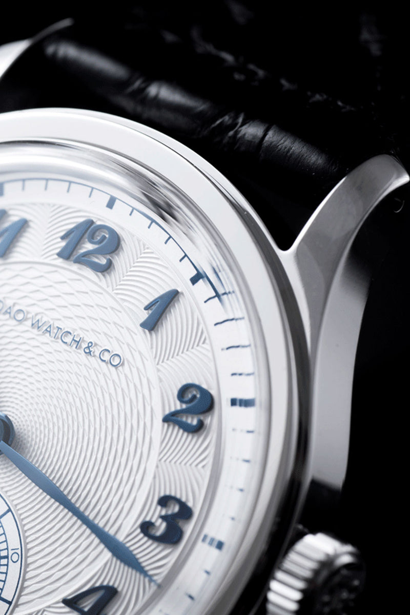 Close-up of a Bao Watch & Co. wristwatch featuring a white textured dial with blue Arabic numerals, a polished silver-tone case, and a black leather strap against a dark background.