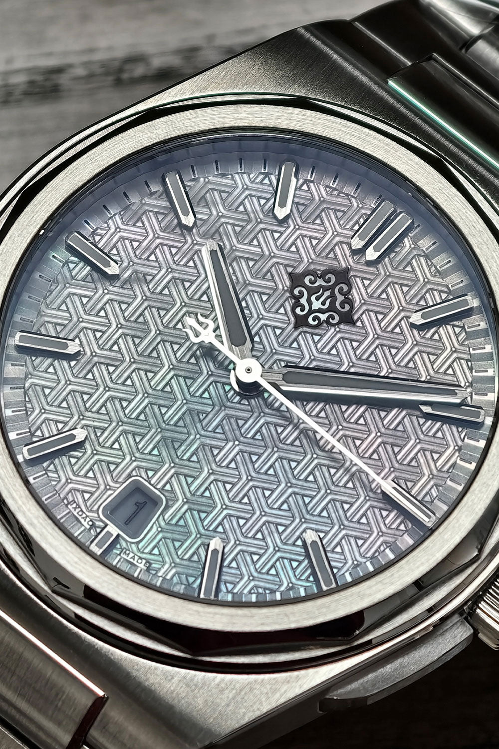 Close-up of a wristwatch with a silver-toned metal bracelet and a textured geometric dial in a blue-gray gradient. The watch has silver hour markers, silver hands, and a date window at the 3 o'clock position.
