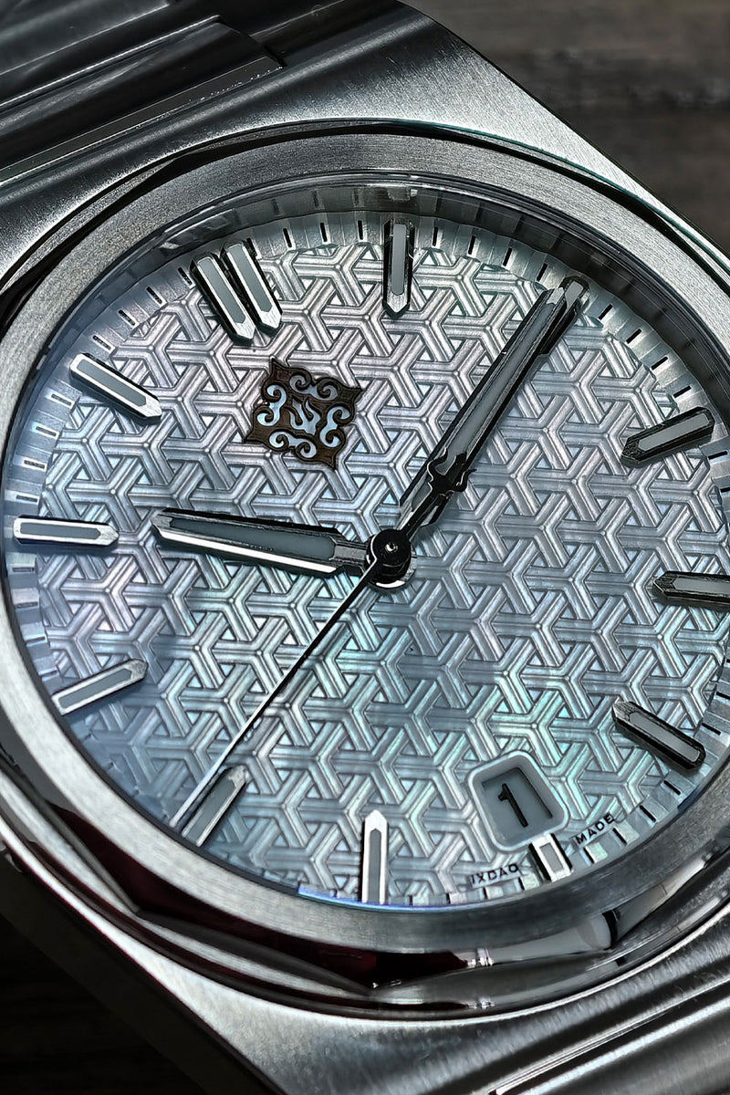 Close-up of a wristwatch with a silver-toned metal case and a textured geometric dial in pale blue-gray tones. The watch has silver hour markers and a date window at the lower right.