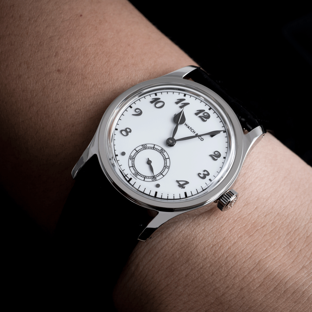 A close-up of a wrist wearing a Vacher-Serfontaine watch with a silver-toned case, white dial, black Arabic numerals, and a black leather strap against a dark background.